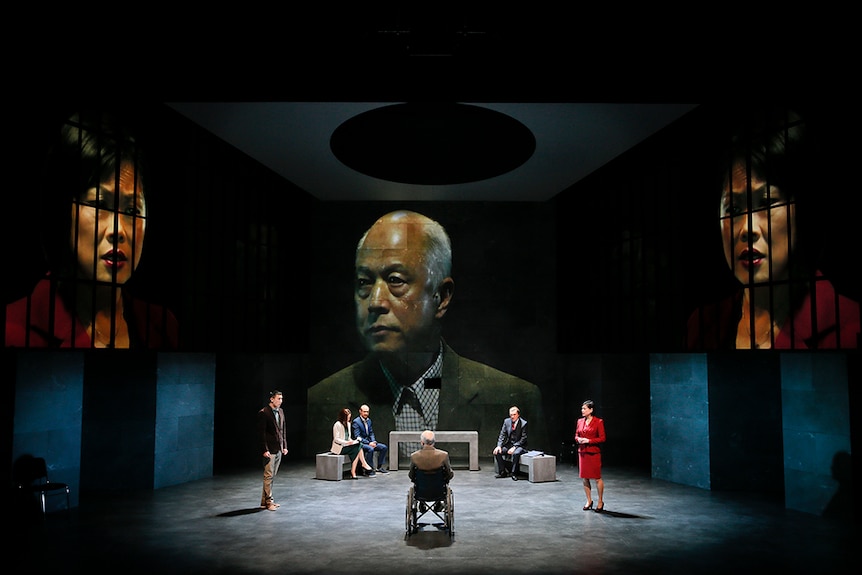 A man in wheelchair with back to audience faces two standing people, three seated on concrete benches and large projections.