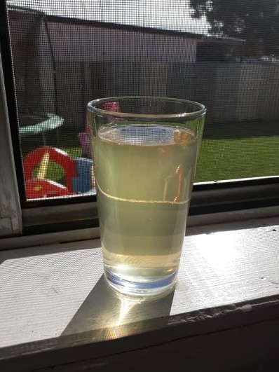 A glass of discolored water sits in a window sill with a playground for children in the background.