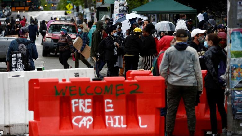 People walk past a barricade that reads' Welcome 2 'Free Cap Hill' spray painted on it in white and green