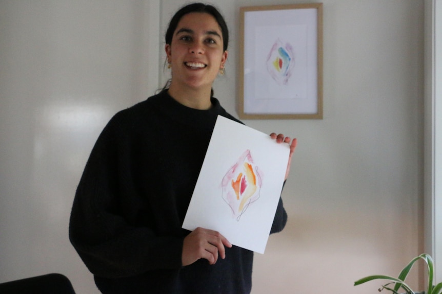 A smiling, dark-haired woman in a dark jumper holds up a small painting of a vagina.