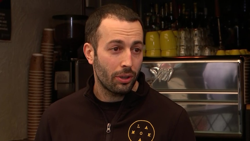 A man with black hair and a beard standing in front of a coffee machine 