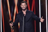 Chris Hemsworth, a white man with blonde hair, wears a sleek black suit and holds an award statue on a stage.