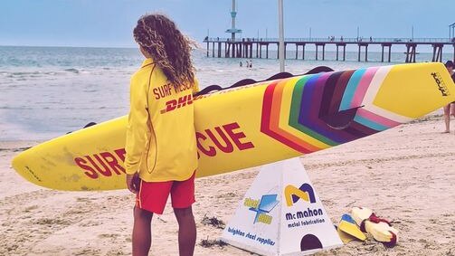 woman lifesaver with her back to the camera on a beach, jetty in the background, holding a surf rescue board