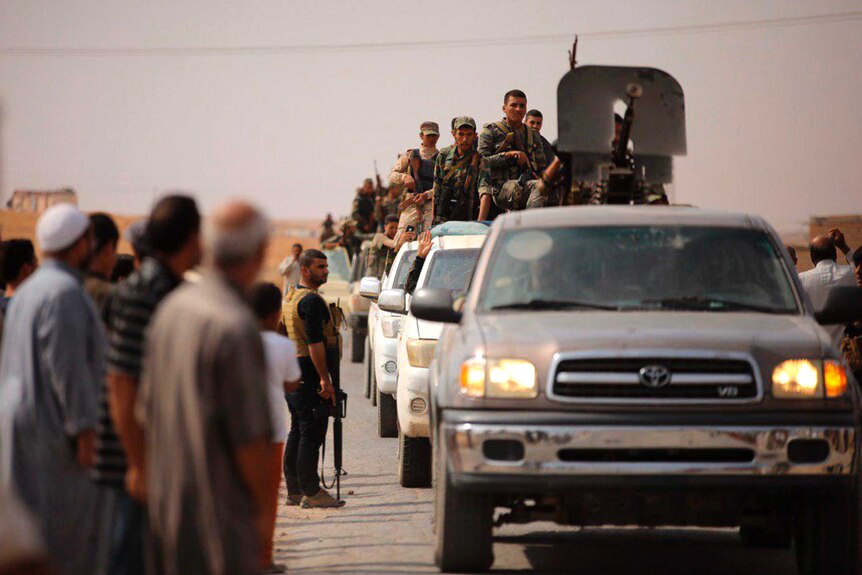 Syrian troops enter the town of Ein Issa in a convey of trucks as civilians stand on the road side.