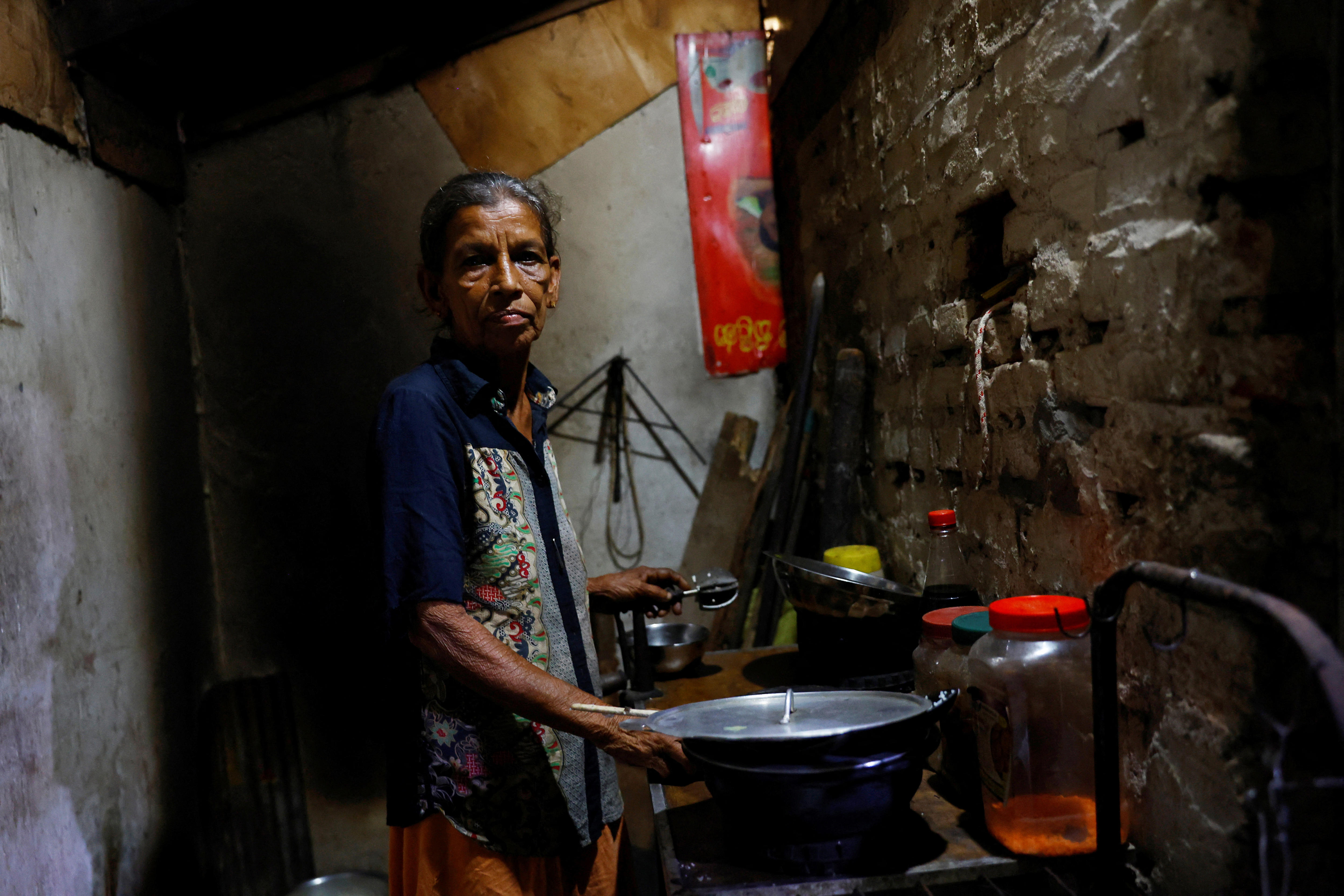 An elderly Sri Lankan woman stands in a small brick kitchen in low light.