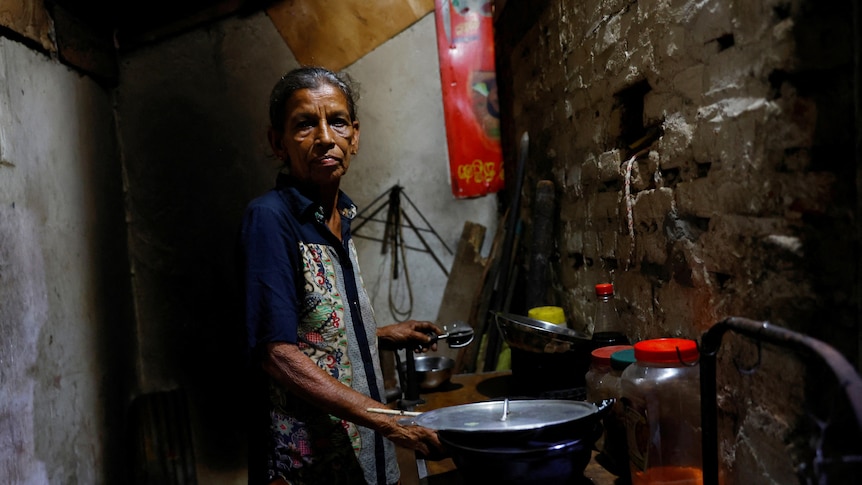 An elderly Sri Lankan woman stands in a small brick kitchen in low light.