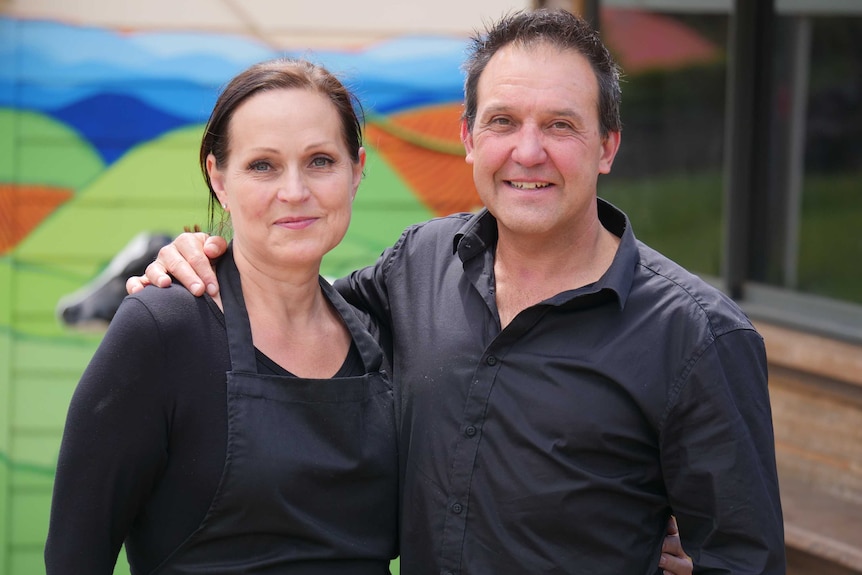 A middle aged man and woman with dark hair stand together with their arms around each other.