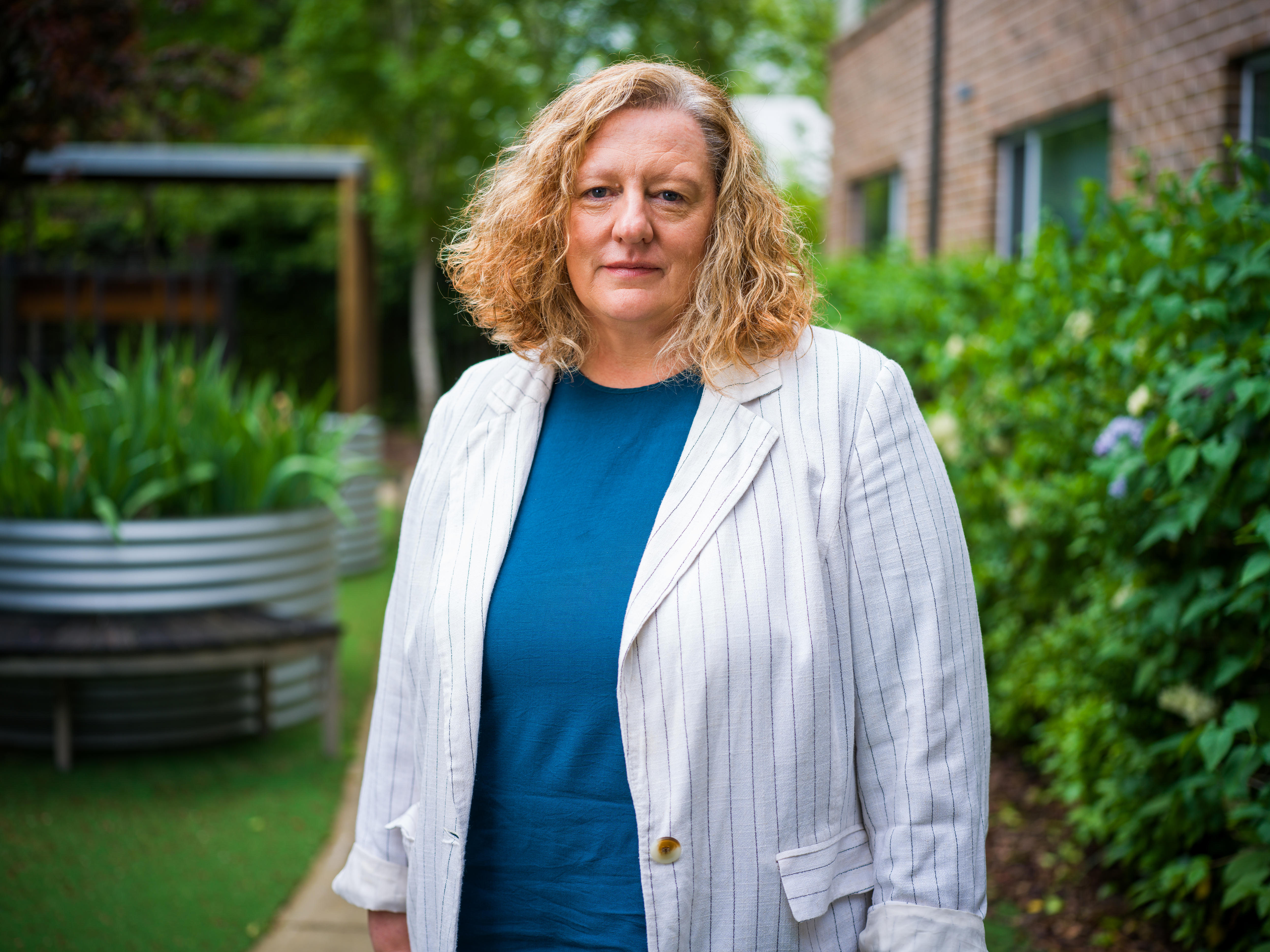 A woman in a white striped blazer and midlength blonde curly hair looks at camera, standing in garden.