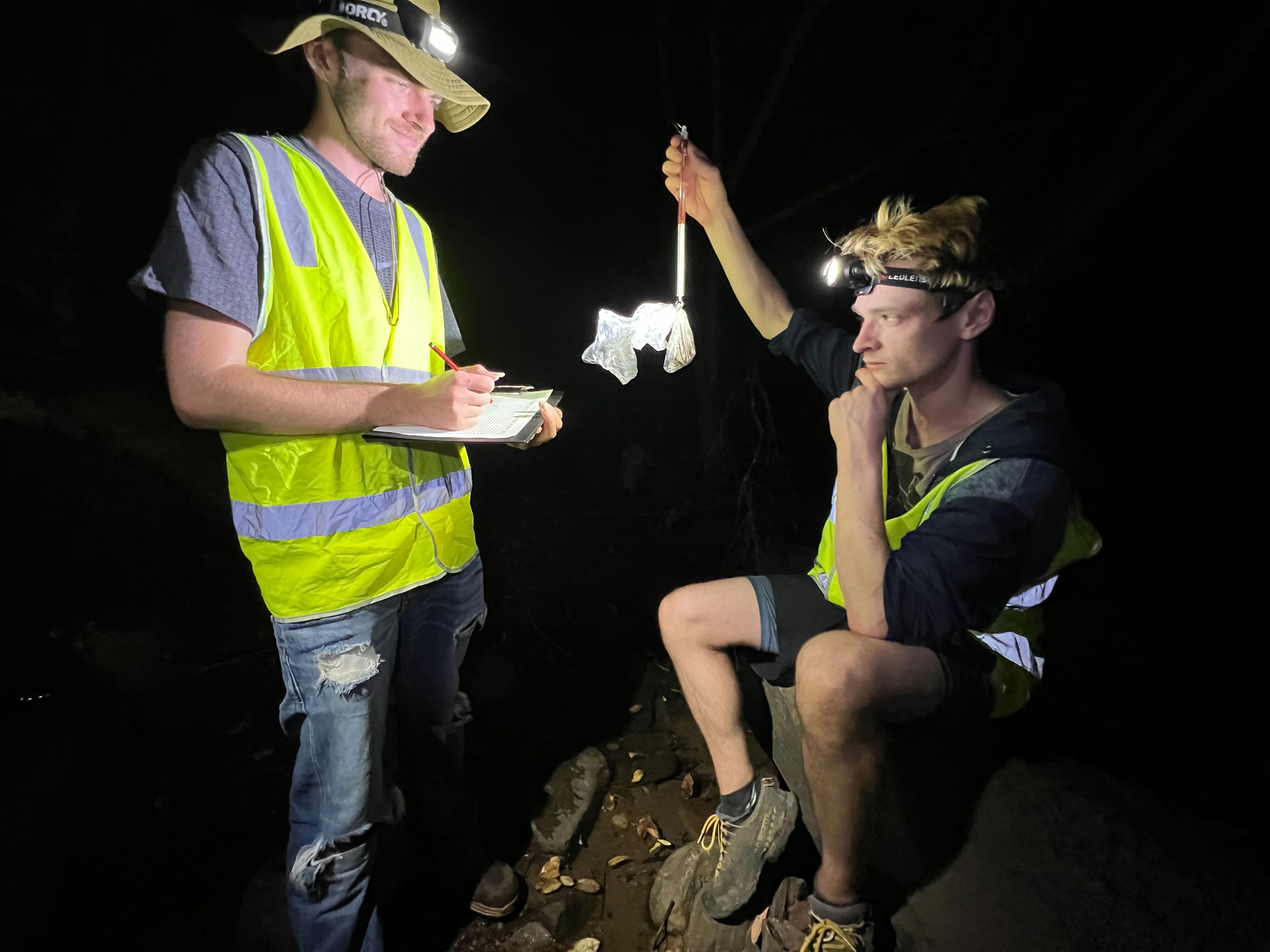 At night a white young man in a hi-vis vest and floppy hat holds a clipboard, a second man holds a bag.