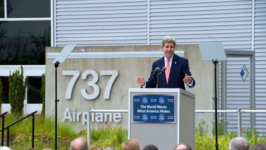 A crowd listens to a Caucasian man speaks on a lectern with the numbers '737' fastened to concrete behind him.