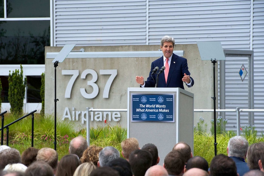 A crowd listens to a Caucasian man speaks on a lectern with the numbers '737' fastened to concrete behind him.