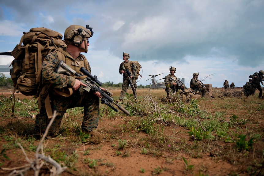 A small group of US Marines crouching or standing in a row in red dirt, holding rifles.