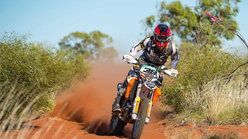 A dirtbike rider stands on his bike, moving fluidly between desert blossoms.