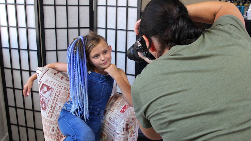 A young girl with long blue dreadlocks is posing on a chair as a photographer takes her photo