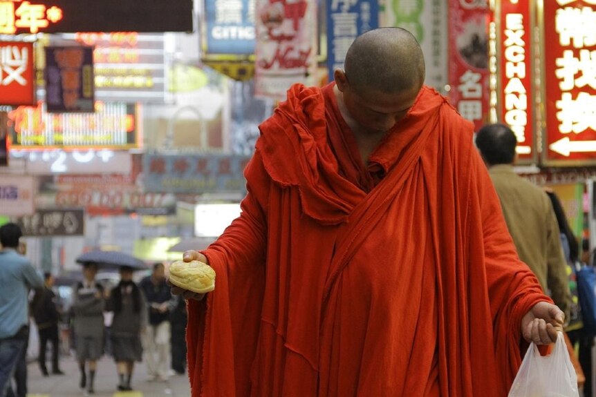 A monk dressed in red orange robes holds plastic bag and pineapple bun in each hand looks at ground in Hong Kong street scene.