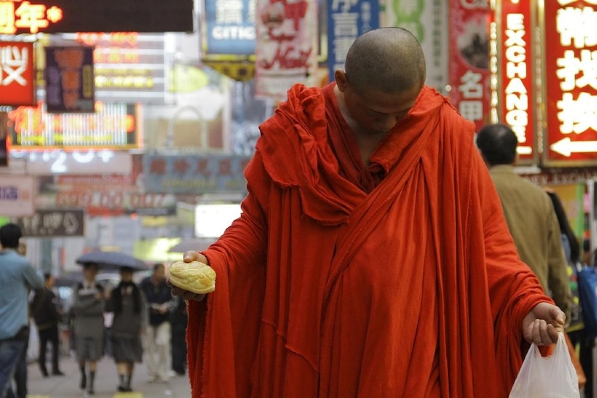 A monk dressed in red orange robes holds plastic bag and pineapple bun in each hand looks at ground in Hong Kong street scene.