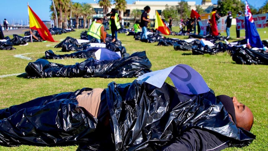 People lie in mock body bags on grass next to St Kilda beach as part of a protest