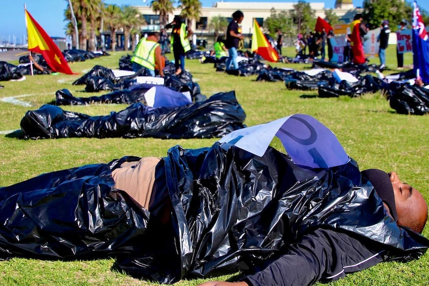 People lie in mock body bags on grass next to St Kilda beach as part of a protest