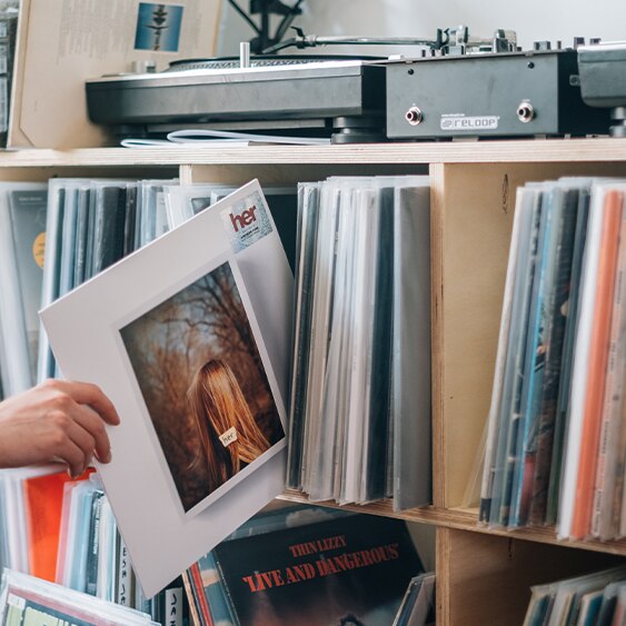 A hand stretches out to grab a record from a collection in a cabinet beneath a record player.