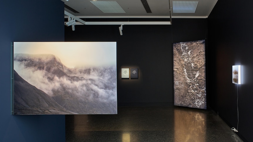 In a gallery, a photograph of an arctic mountain landscape juts out from the wall. Behind it is a landslide image.