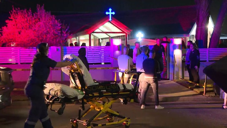 A paramedic rolls a stretcher towards a church while a crowd stand outside.