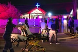 A paramedic rolls a stretcher towards a church while a crowd stand outside.