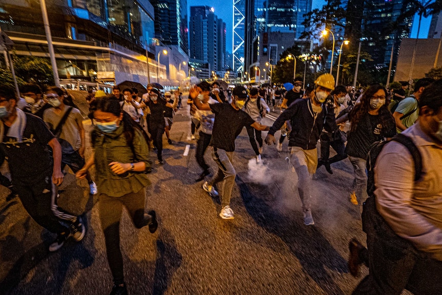 People are blurry as they run on a street, some smoke from tear gas can be seen rising, they wear hard hats, surgical face masks