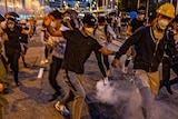 People are blurry as they run on a street, some smoke from tear gas can be seen rising, they wear hard hats, surgical face masks