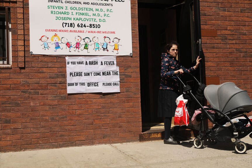 A woman with a pram leaves a building. a sign reads 'if you have a rash or fever please don't come near the door of this office'
