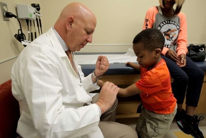 A doctor sits at a chair evaluating the stumps of a young boy's forearms.