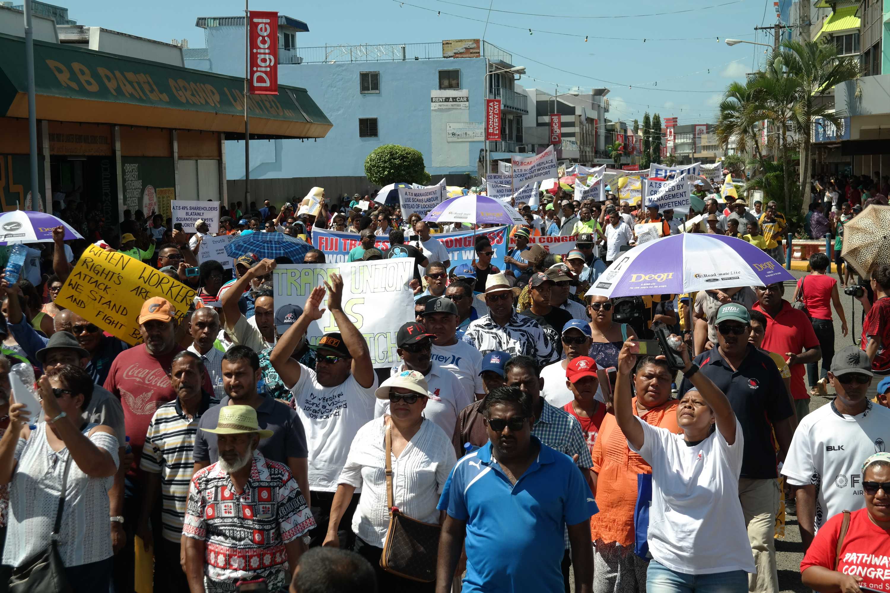 A crowd of protestors walks down a Nadi street, holding signs and clapping.