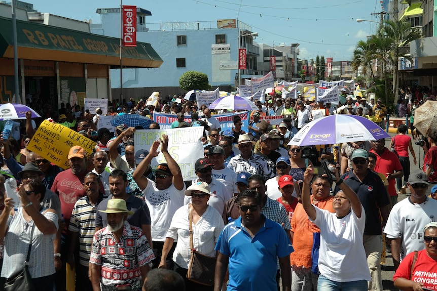 A crowd of protestors walks down a Nadi street, holding signs and clapping.