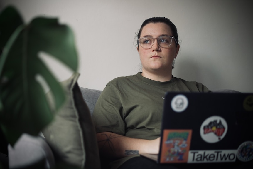 A young white woman with brown hair and glasses. She is sitting on a couch with a laptop