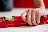 A man's hand holds a red spirit level even on a white tiled surface.