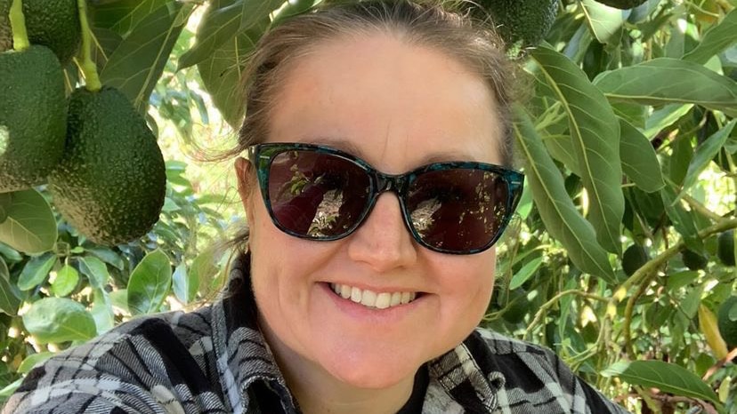 A smiling woman wearing sunglasses stands under an avocado tree. 