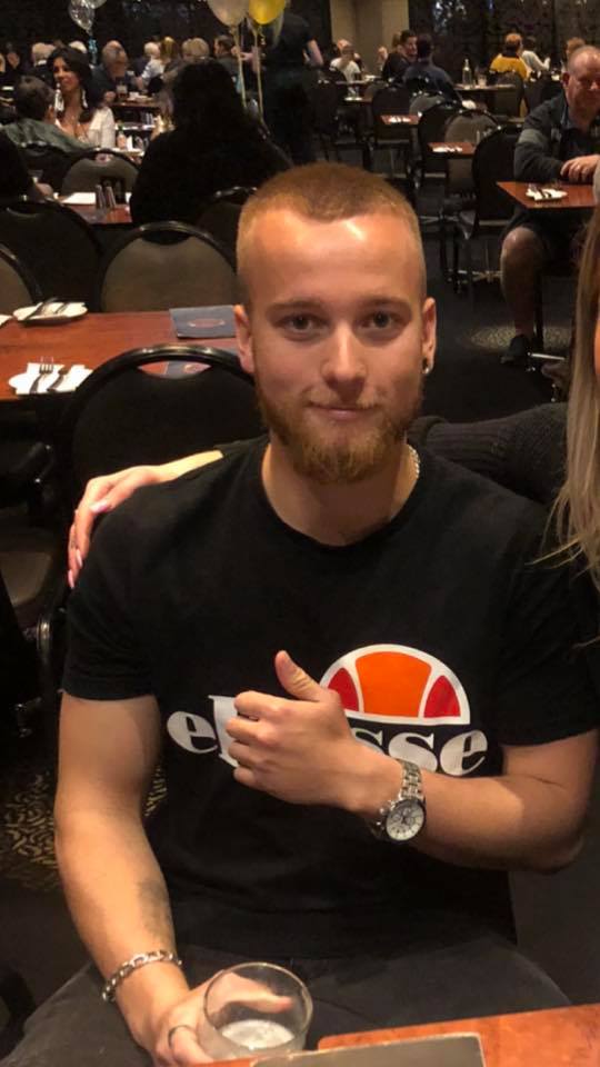 A young man sits at a restaurant table, smiling and giving a thumbs up. Someone off frame has her arm around him.