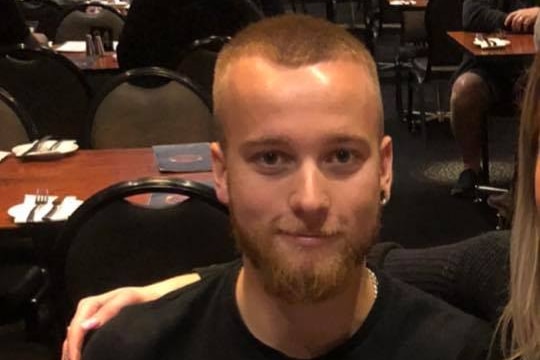 A young man sits at a restaurant table, smiling and giving a thumbs up. Someone off frame has her arm around him.