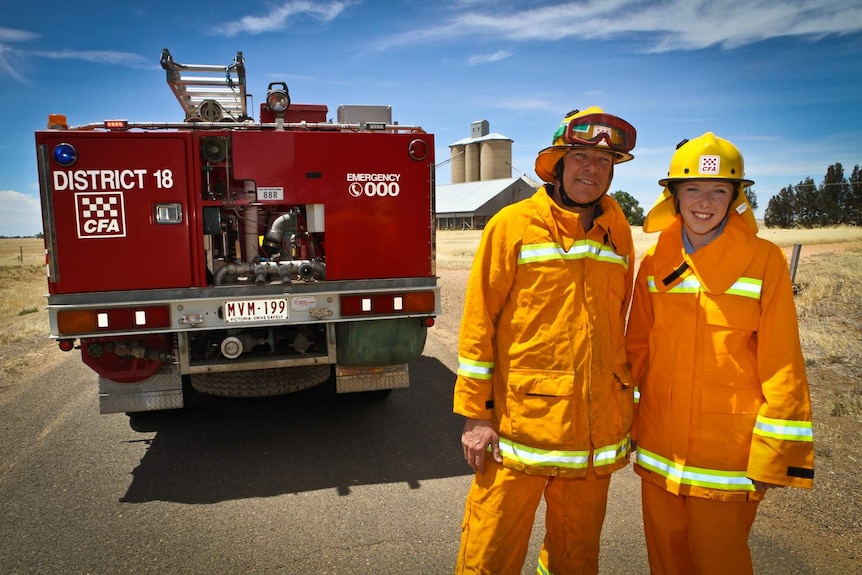 A man and young woman in yellow overalls stand beside a fire truck