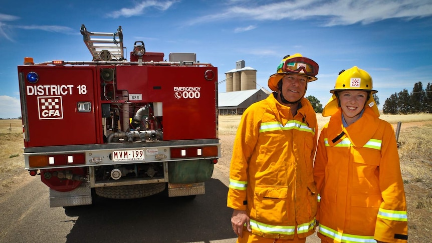 A man and young woman in yellow overalls stand beside a fire truck