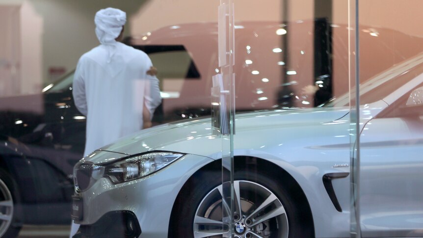 a man in a kandura stands inside a car dalership surrounded by luxury cars