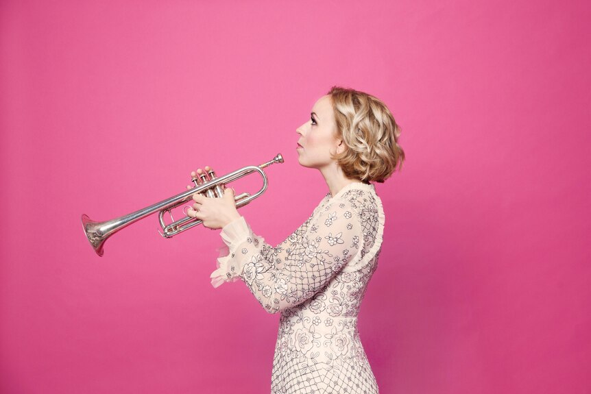 A woman stands side on holding a trumpet. She wears a cream coloured dress and stands against a bright pink wall.