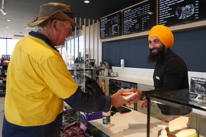 Indian man on right behind counter passing take-away container to man with hat inside roadhouse shop.