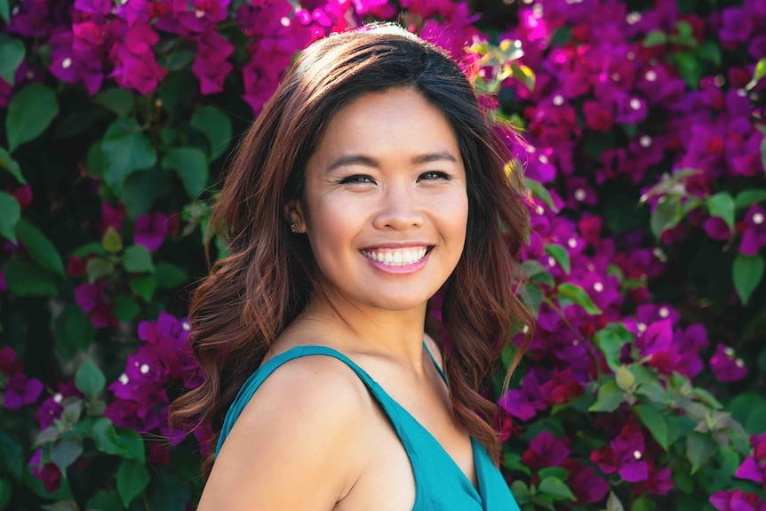 A young woman in a teal jumpsuit with magenta flowers in the background.