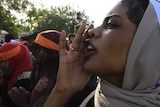 A close-up image of a woman in a brown hijab and with a white sash calls out with her hand in a large crowd.