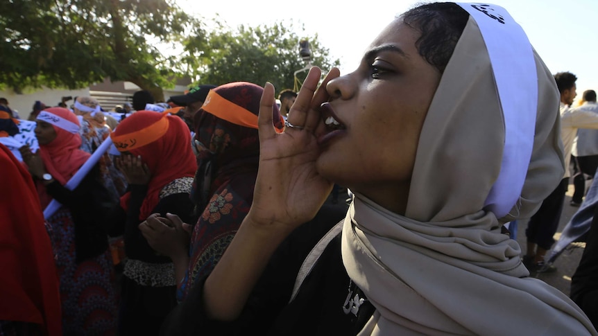 A close-up image of a woman in a brown hijab and with a white sash calls out with her hand in a large crowd.