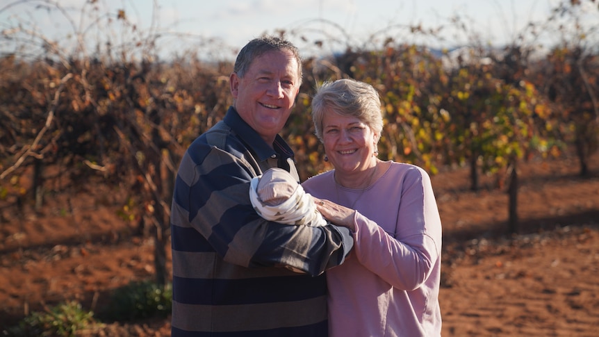 A man and woman standing in front of a vineyard holding their foster baby