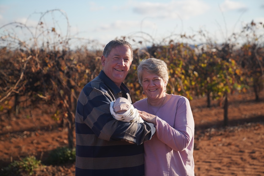 A man and woman standing in front of a vineyard holding their foster baby