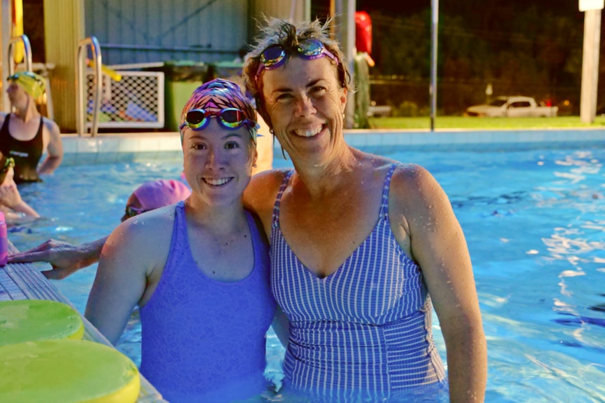a woman stands with her hand around a younger woman while in a swimming pool at night time 