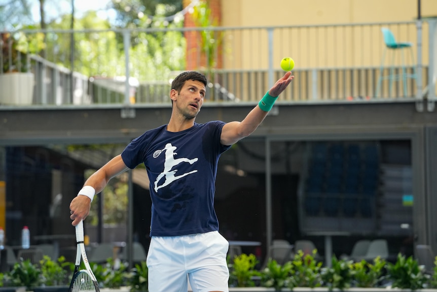 A man wearing a blue shirt and white shorts holds a tennis racquet in one hand and lifts a ball up with the other