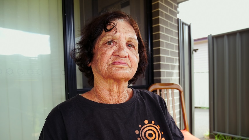 Woman sits in black t-shirt with Aboriginal art logo, in front of grey brick house with glass door.